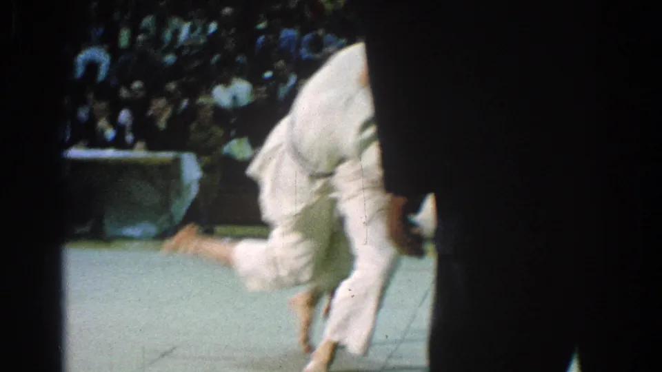During the San Gabriel judo match, a competitor in a white gi bends or falls near the mat as an official partly blocks the frame.