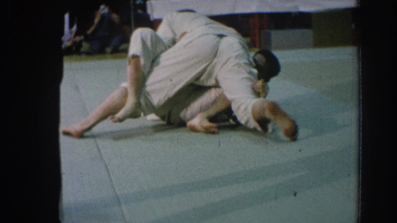 Two martial arts competitors in white gi grapple on a pale mat.