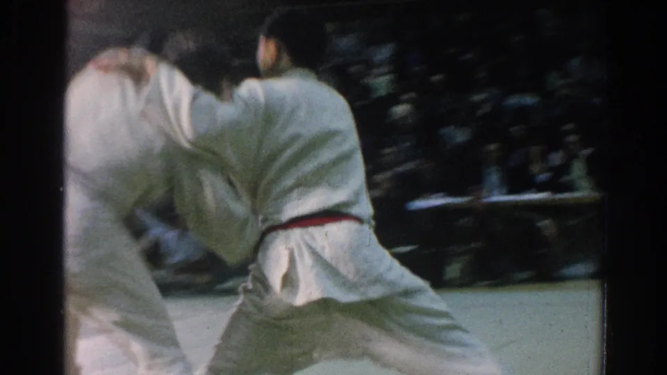 Two white-gi competitors appear upright in a fast exchange, with blurred movement and seated spectators behind them.