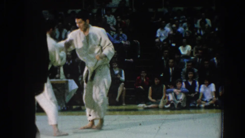 A white-gi competitor stands during the 1966 San Gabriel match while spectators watch from the gym seating area.