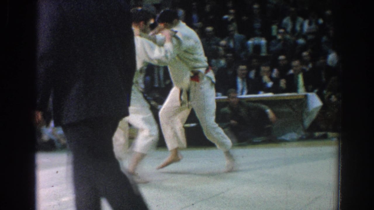 Two men in white gi grapple on a mat while a suited official stands nearby.