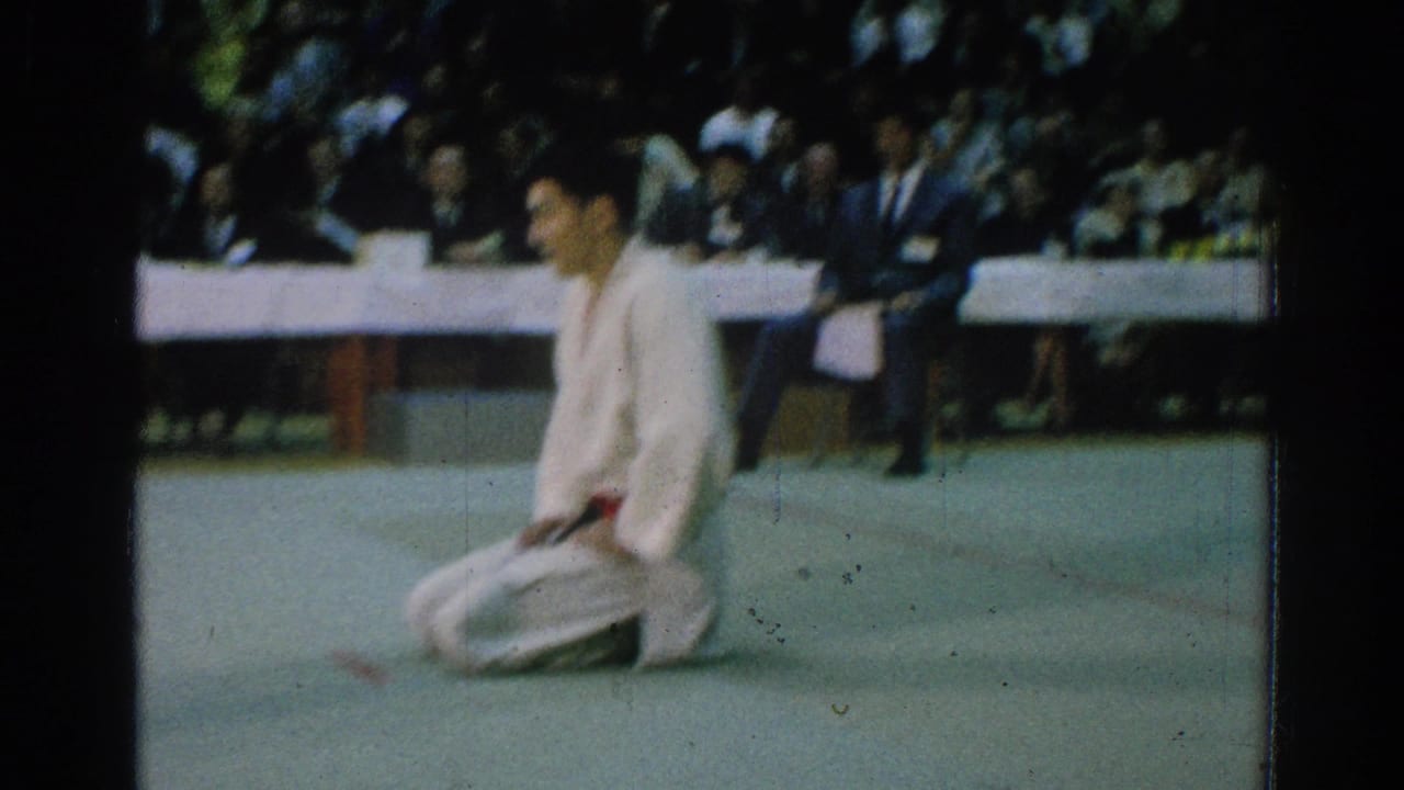A competitor in a white gi kneels on the mat with spectators behind him.