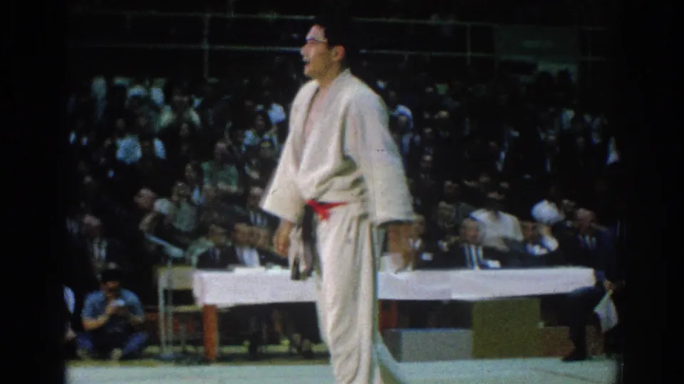 A competitor in a white gi with a red belt stands on the mat in the 1966 San Gabriel judo footage, with spectators and a table behind him.