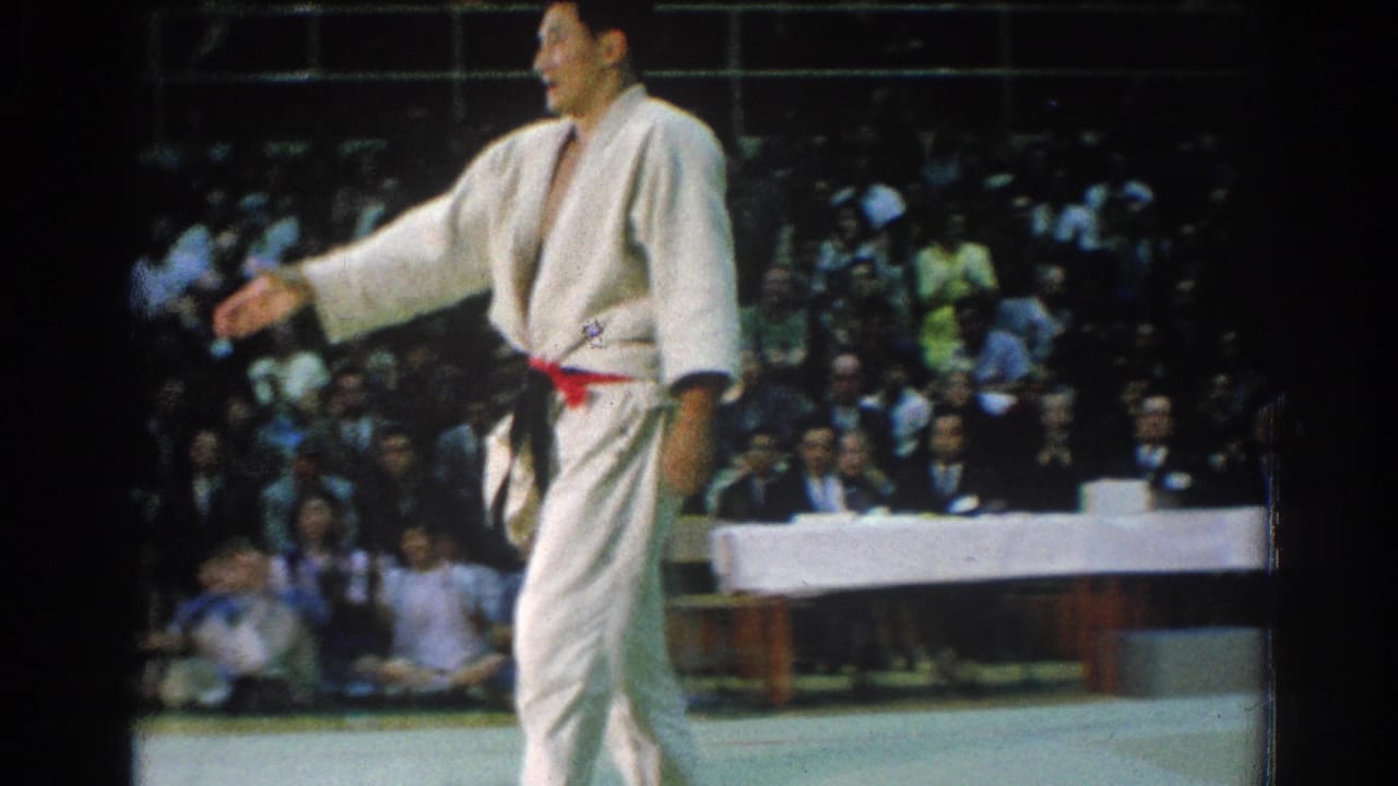 A karate competitor in a white gi with a red belt stands in profile during a 1966 tournament in San Gabriel, California, with a seated crowd visible behind the mat.