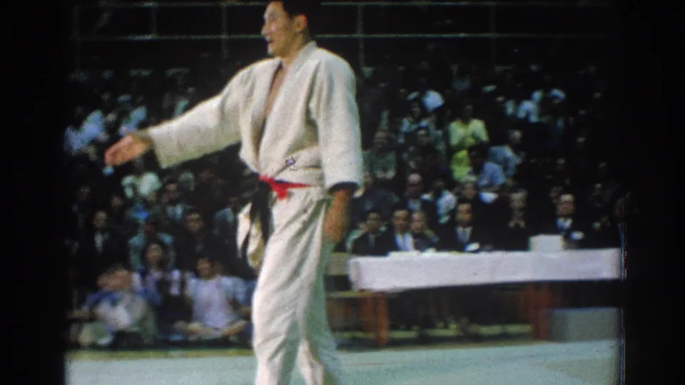 A karate competitor in a white gi with a red belt stands in profile during a 1966 tournament in San Gabriel, California, with a seated crowd visible behind the mat.