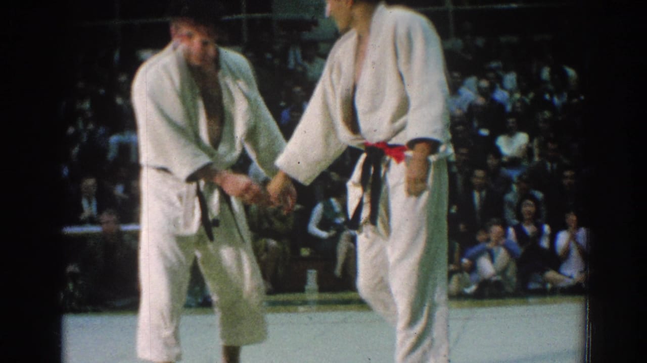 Two karate competitors in white gi shake hands during the San Gabriel tournament, with spectators and officials seated in the background.