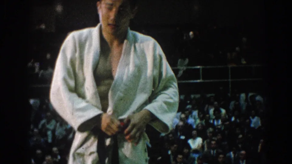 A young male karate competitor adjusts his belt or gi in a close home-movie view, with the tournament crowd blurred behind him.