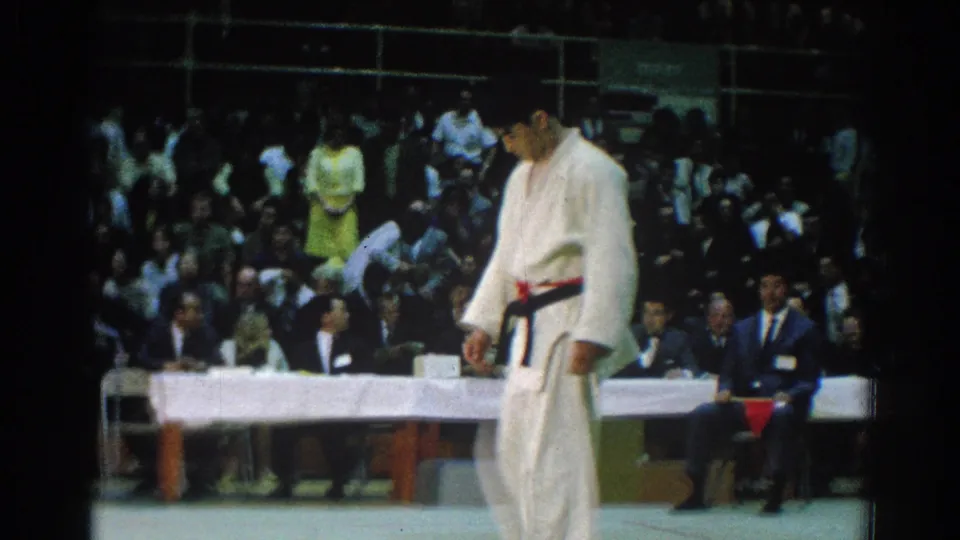 A karate competitor in a white gi and red belt lowers his head near the judges' table during the 1966 San Gabriel tournament, with spectators filling the background.