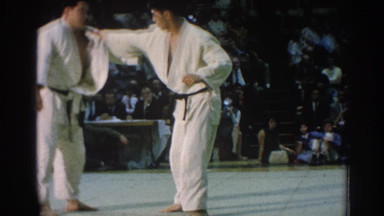 Two martial artists in white gi face each other on a mat before seated spectators.