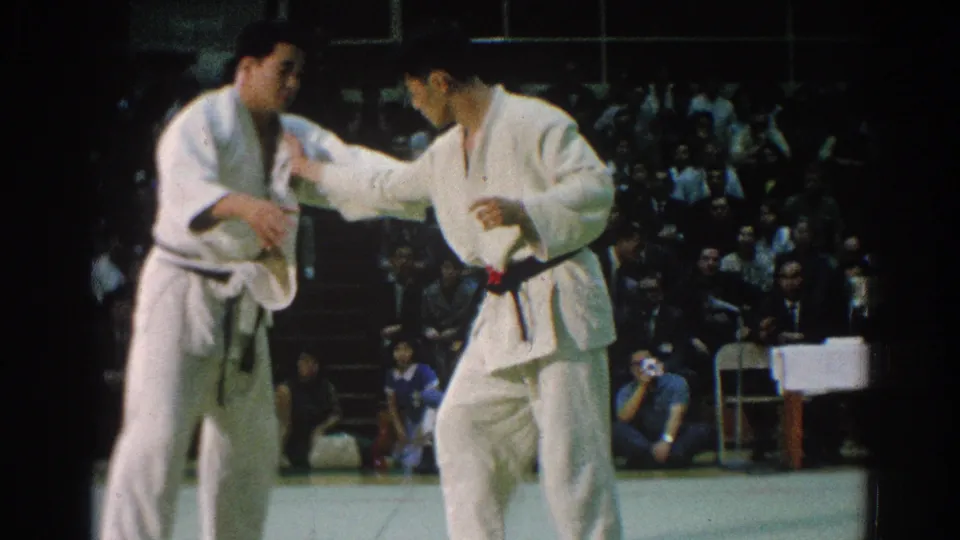 Two martial artists in white gi stand during the San Gabriel tournament footage, with the audience and chairs behind the mat.