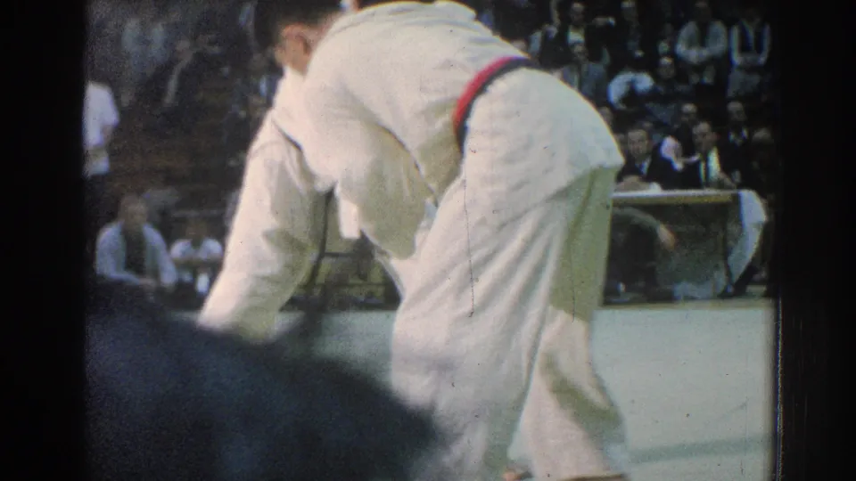 A close, partially obstructed 1966 San Gabriel frame shows a martial artist bent forward as the match continues near the audience.