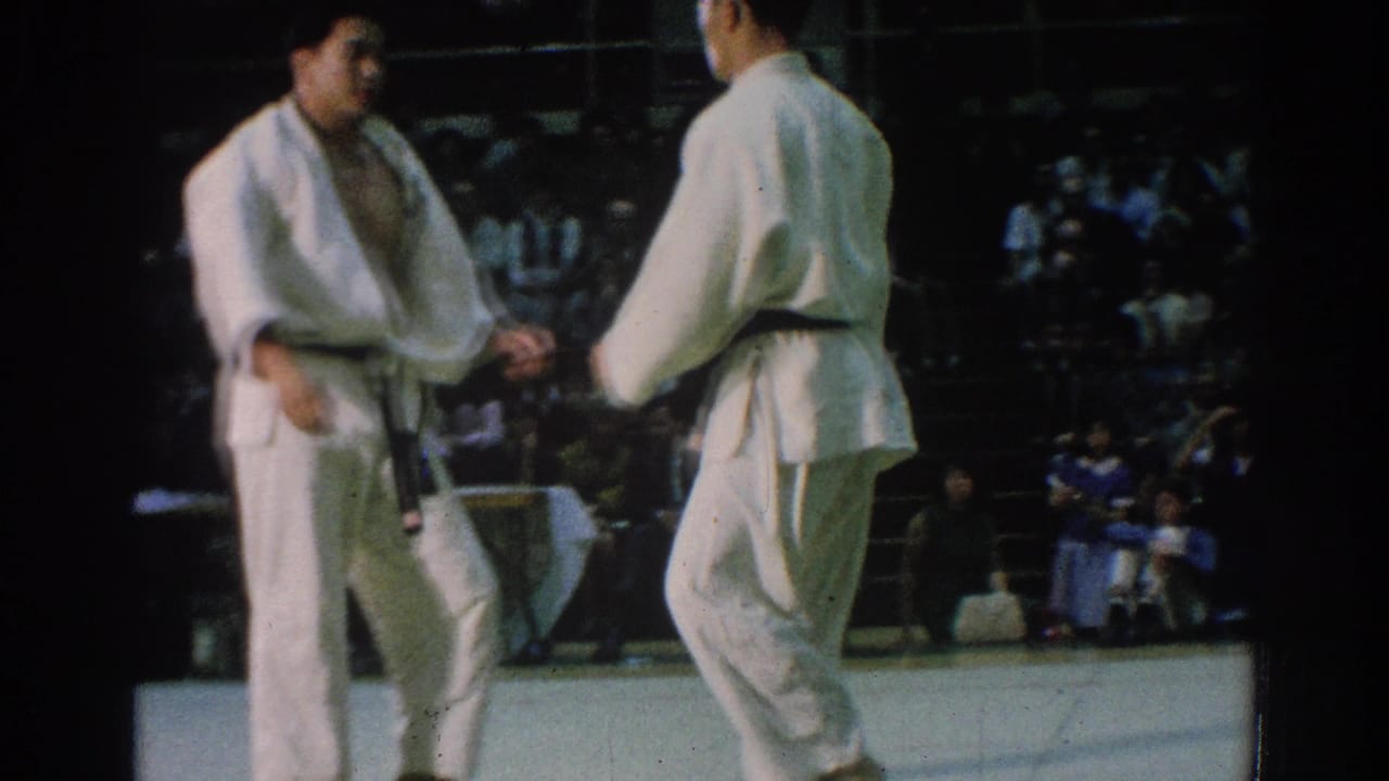 Two judo athletes in white gi face each other on a mat with spectators behind them.
