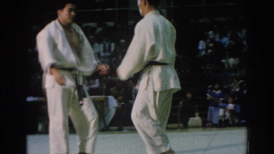 In San Gabriel, California in 1966, two judo athletes in white gi and black belts face each other before a match in a crowded indoor arena.