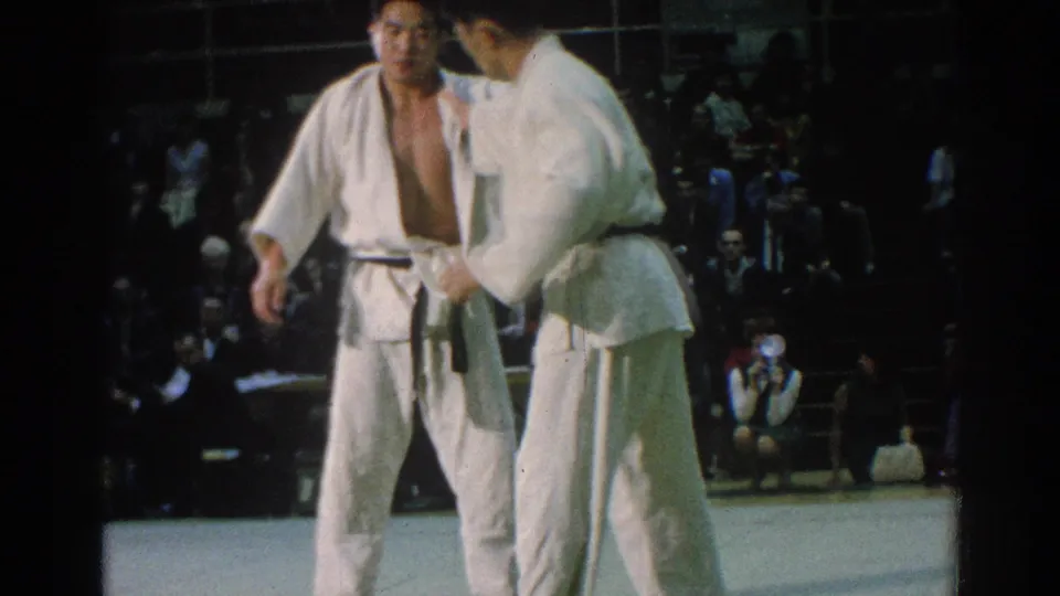 Two black-belt judo athletes stand close on the competition mat in 1966 San Gabriel footage, with one checking or adjusting the other's gi.