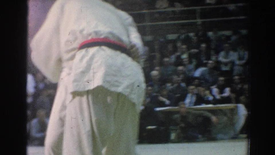 A close, partially cropped 1966 San Gabriel frame shows a judo athlete's white gi and dark belt against the blurred arena audience.