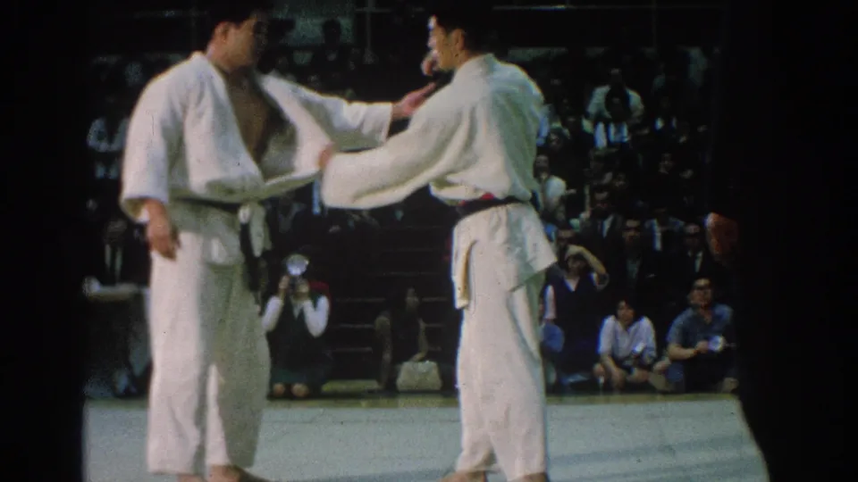San Gabriel 1966 judo competitors in white gi stand on the mat as one reaches toward the other's uniform.