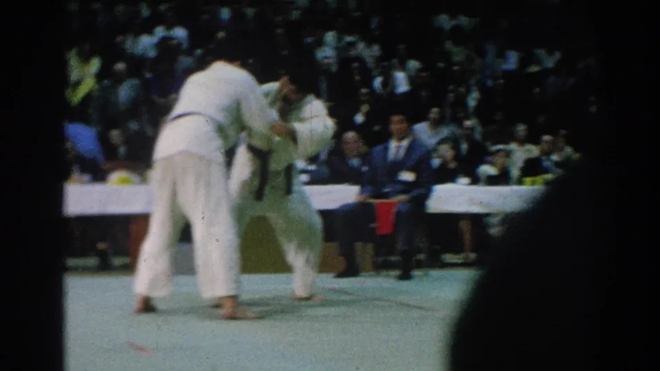Two judo athletes in San Gabriel competition footage move into a grip while spectators and a suited official watch from behind.