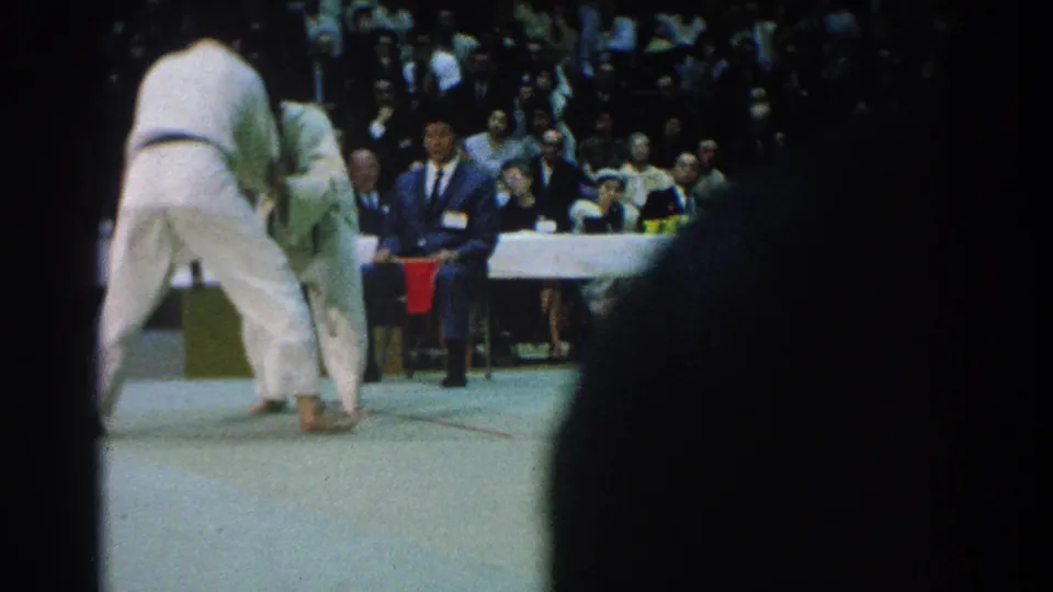 A rear view of a judo competitor shows the pair locked in close grips as a suited official sits near the mat edge.