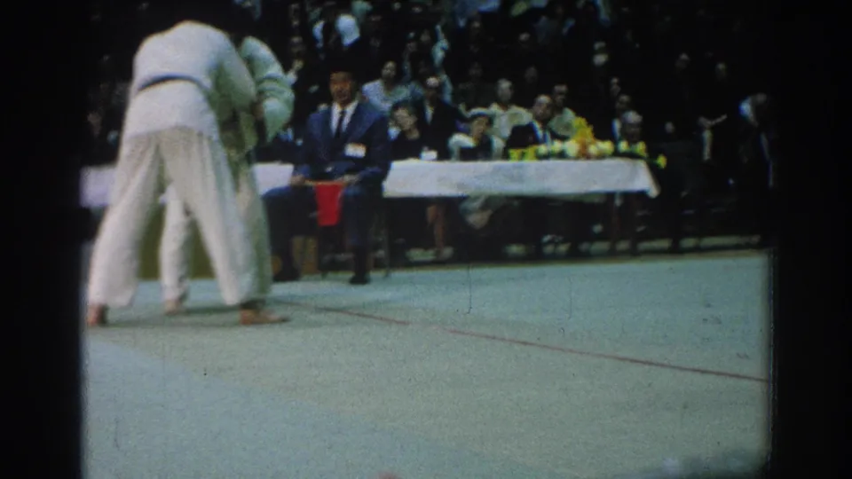 The San Gabriel judo match continues with two athletes in white gi engaged on the mat before a crowded spectator area.