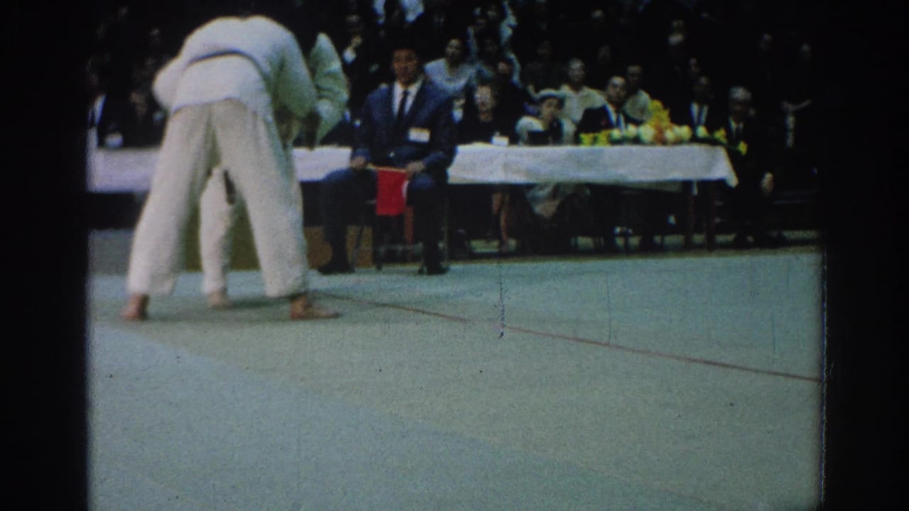 Two competitors in white gi grapple on a mat with a judge seated near a red flag.