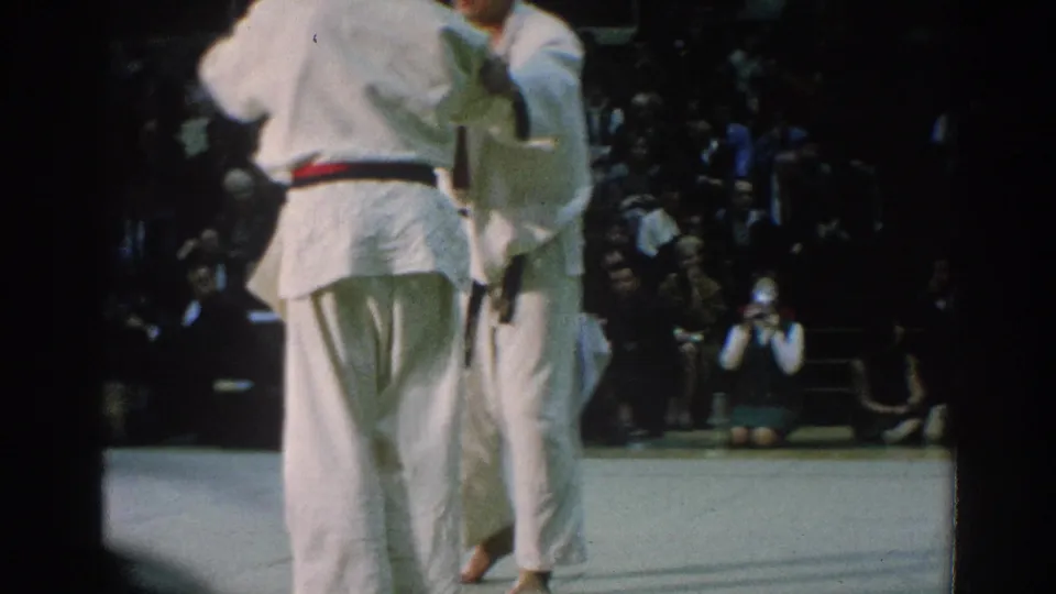 A rear view shows one competitor with a dark belt facing another white-gi fighter, with spectators watching from the dim background.