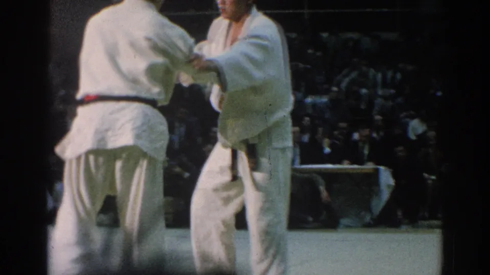 Two competitors in white gi hold sleeve grips during the match, seen from a close spectator angle with the crowd in the background.