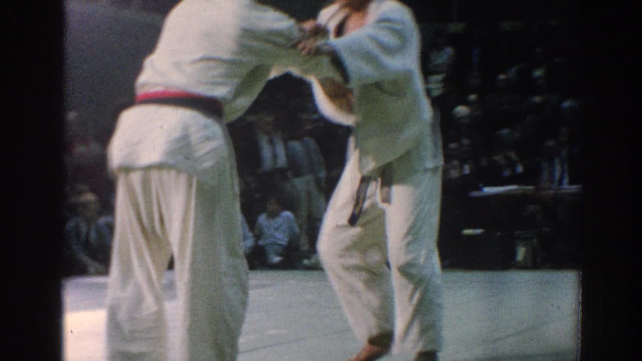 In supported 1966 San Gabriel footage, two gi-clad martial artists grip each other during a match as spectators sit behind them.