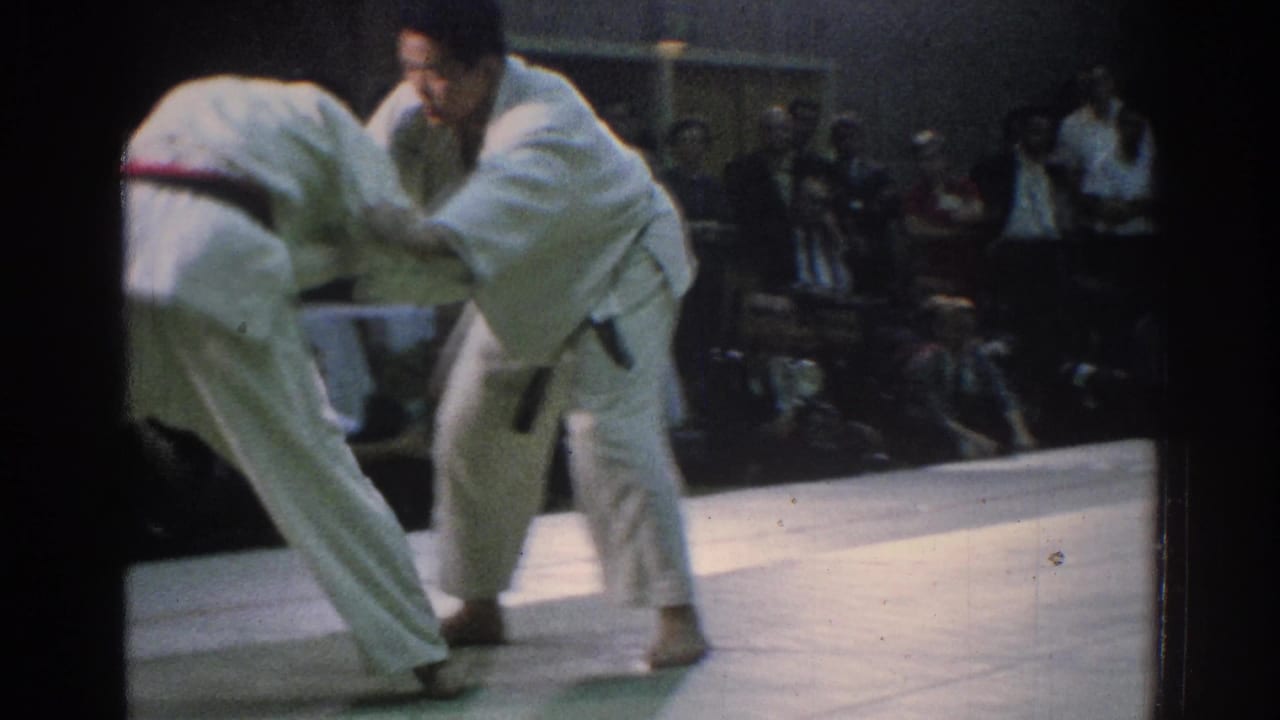 Two martial artists lean into a grapple on a mat with spectators in the background.