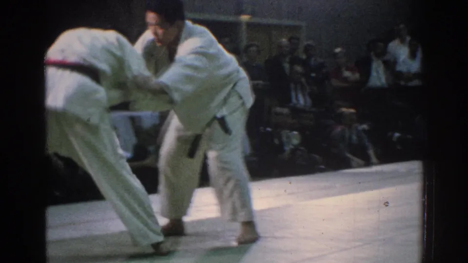 The 1966 San Gabriel home-movie frame shows one martial artist bending forward as the other holds position during a standing exchange.