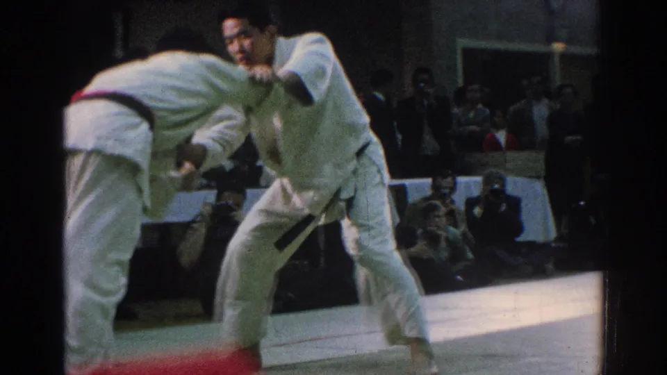 A competitor in a white gi faces toward the camera during a close grapple, with spectators seated beyond the mat in San Gabriel footage.
