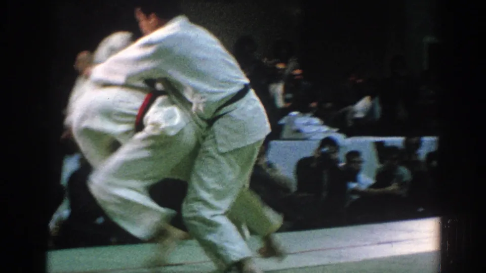 In San Gabriel, California in 1966, two adult men in white judo uniforms grapple during a match as spectators watch from the background.