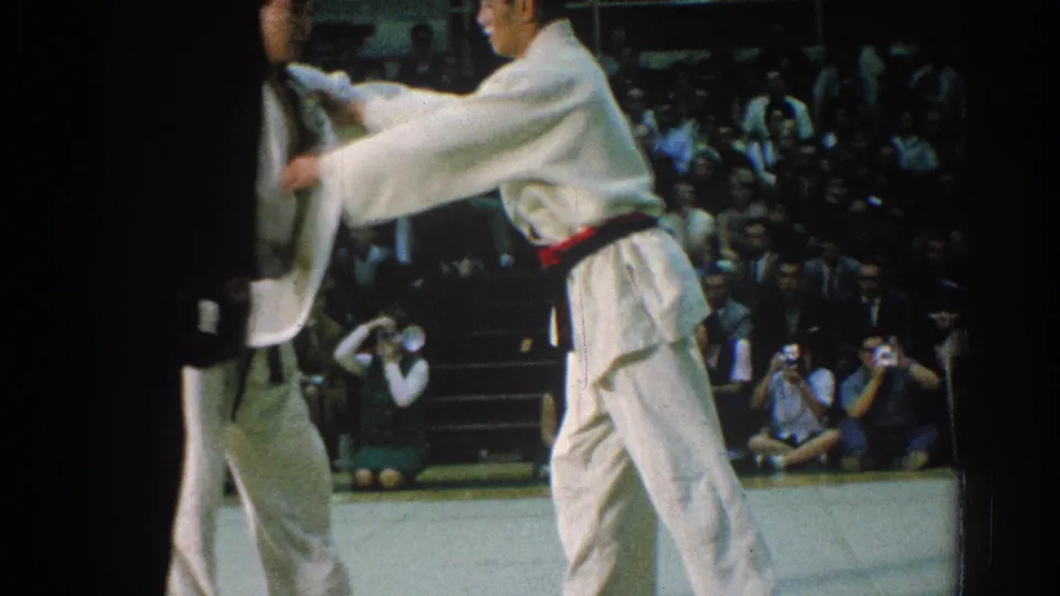 A 1966 San Gabriel judo match frame shows one competitor gripping his opponent's gi near the mat edge, with spectators and cameras behind them.