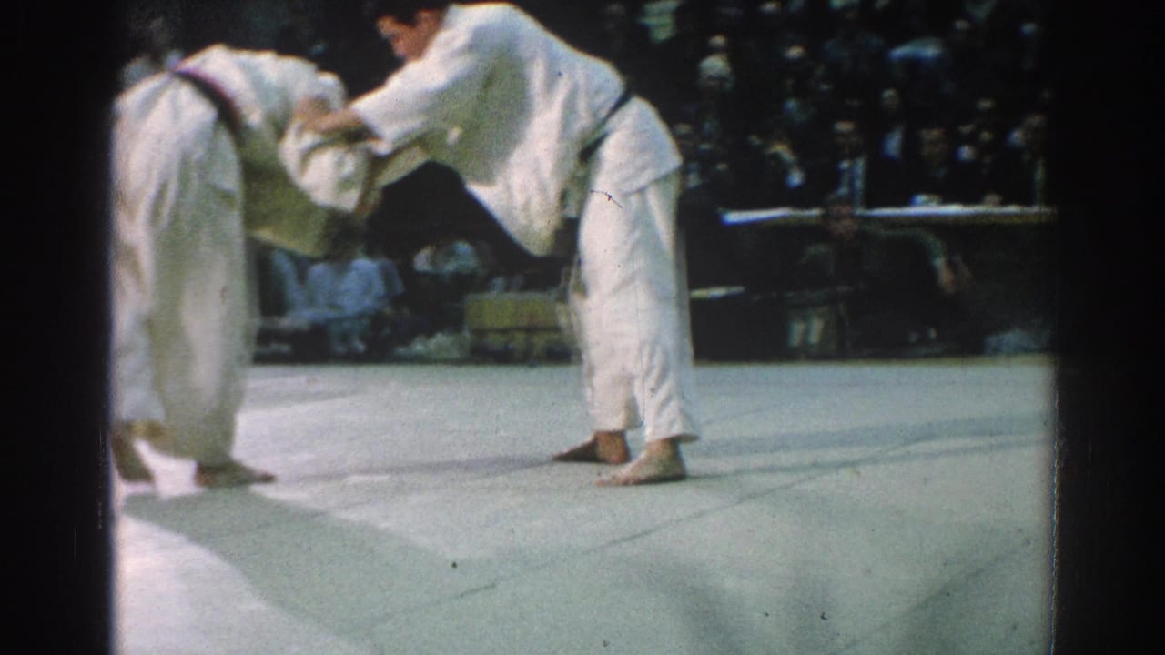Two judo athletes in white gi grip each other on an indoor mat before a crowd.