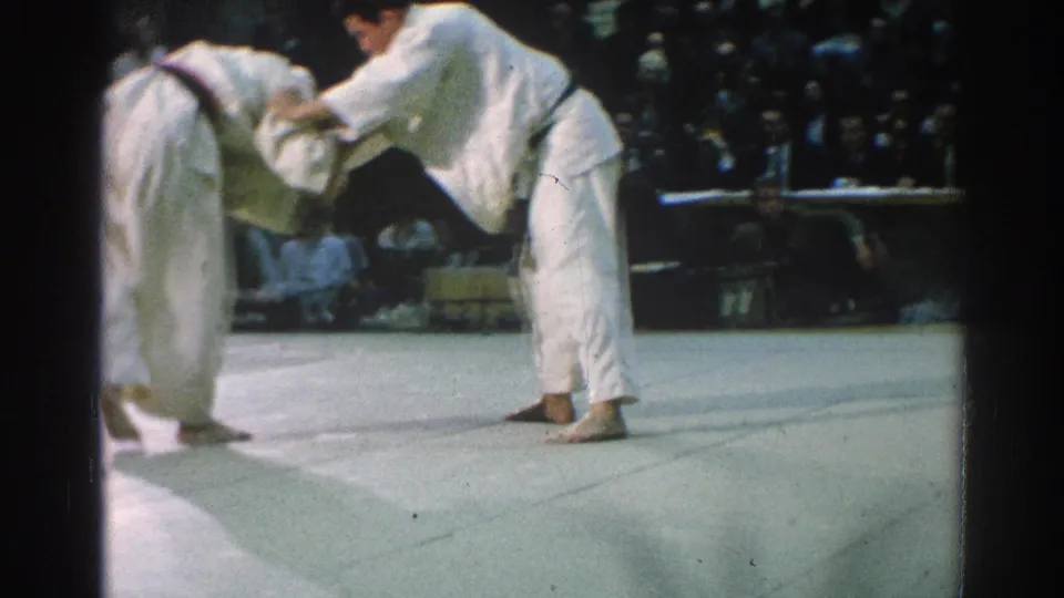 1966 San Gabriel judo competitors in white gi face off on a mat, with spectators visible in the dim background.