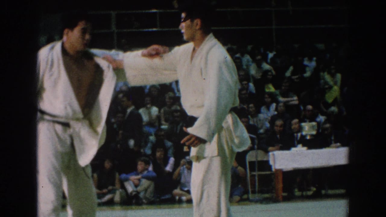 Judo competitors pause at close range while spectators watch from bleachers.