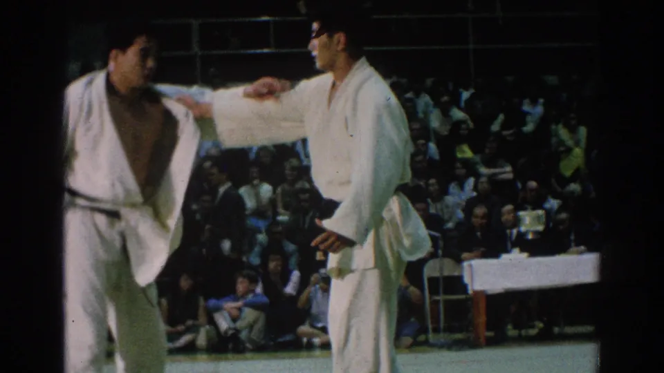 A 1966 San Gabriel judo match shows one athlete extending an arm toward another near the edge of the mat, with audience seating behind them.