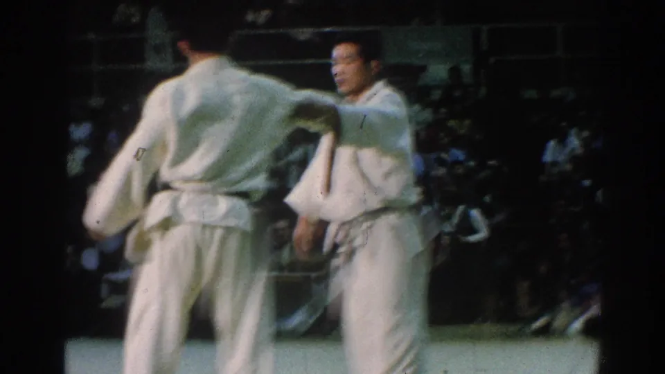 San Gabriel 1966 indoor tournament frame with one competitor facing camera and another seen from behind during a pause or reset.