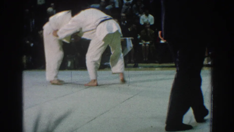 1966 San Gabriel home-movie frame of two martial arts competitors bent into a grapple as a nearby figure watches from the mat edge.