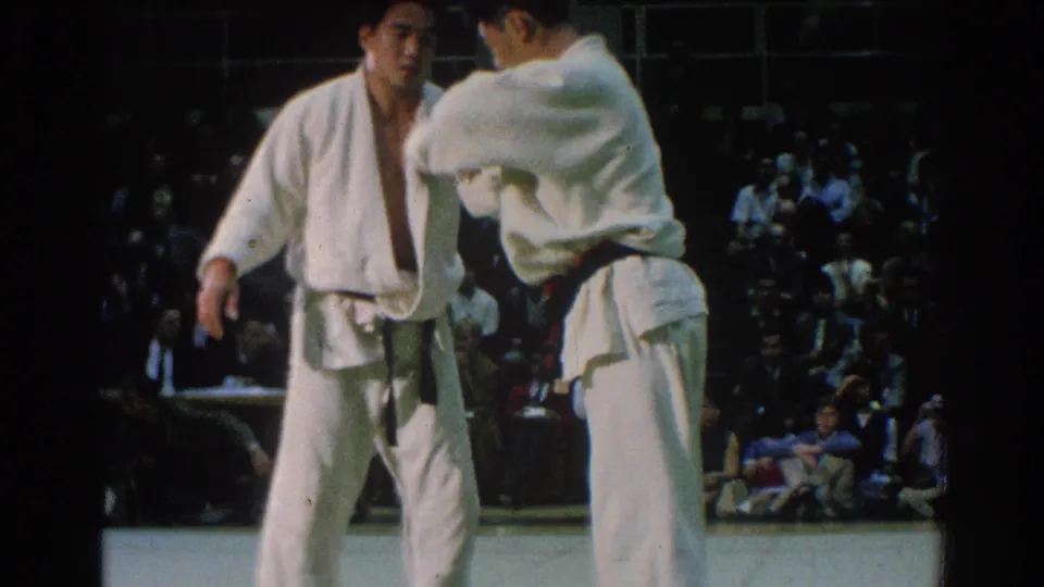 Two competitors in white gi grapple during a 1966 martial arts match in San Gabriel, with a seated crowd in the background.