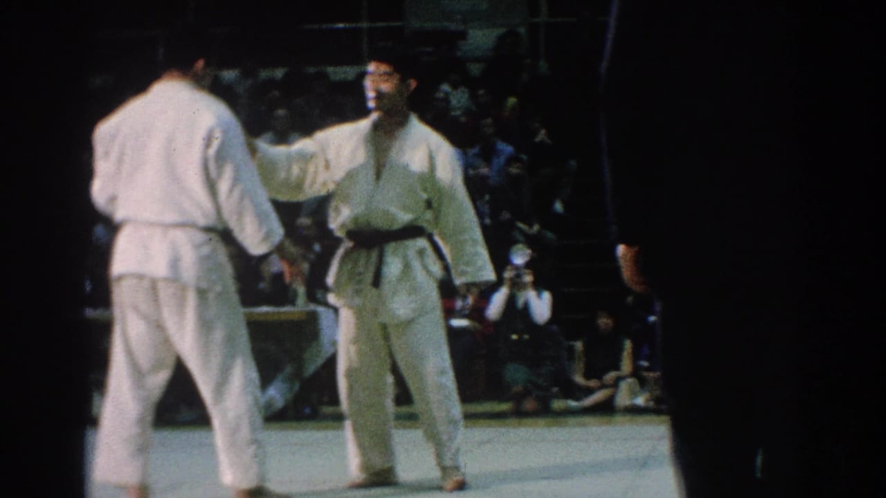Two gi-clad competitors face each other near a referee and seated crowd.