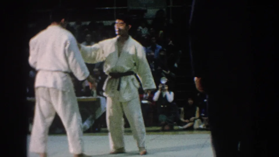 Two martial arts competitors stand on the mat during the San Gabriel match, with an official and spectators visible behind them.