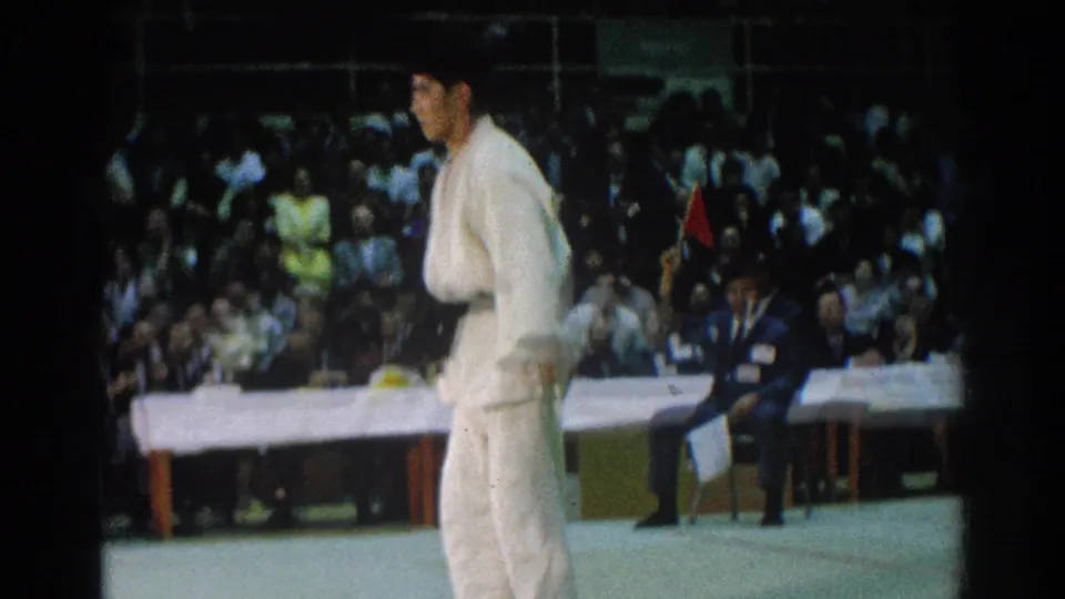 A man in a white gi moves across the mat during the 1966 San Gabriel tournament, with seated spectators and a table behind him.