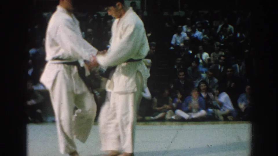 Two martial arts competitors shake hands during the 1966 San Gabriel match, with spectators watching from the background.