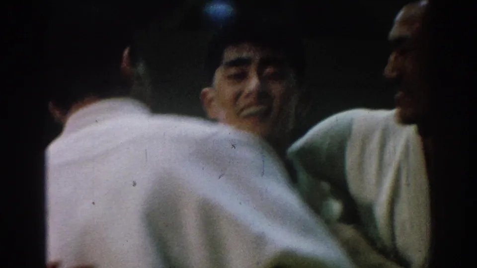 Men in white gi cluster closely during a San Gabriel martial arts contest, with one face visible in the dim frame.