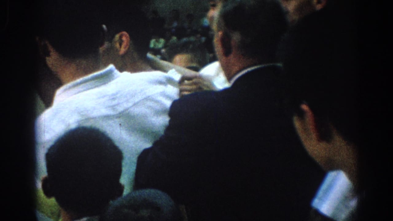 Crowded indoor scene with men seen from behind and a hand pointing across the group.