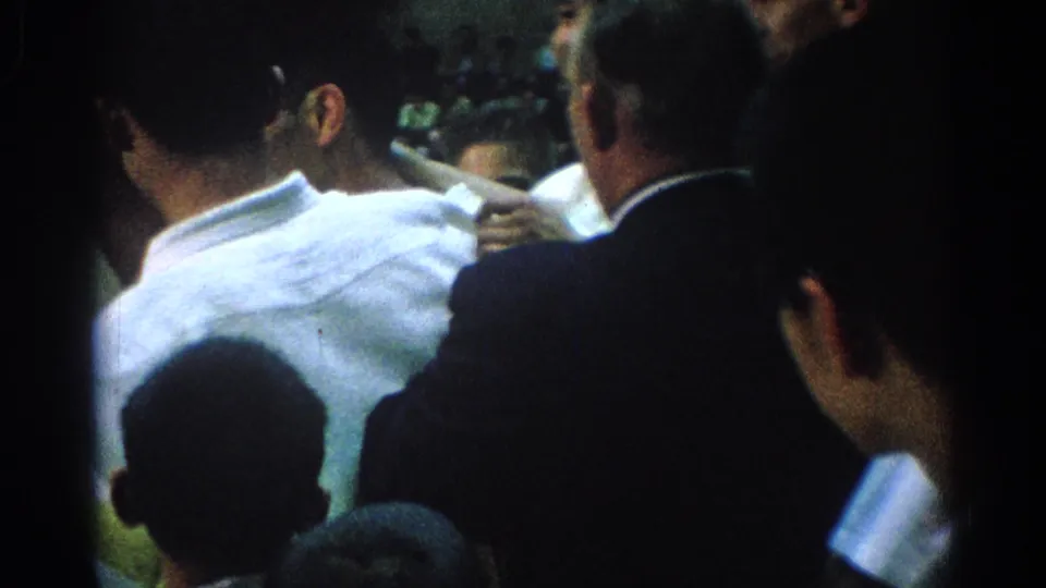 1966 San Gabriel home-movie frame showing a dense indoor crowd with men in white shirts and dark suits viewed from behind.
