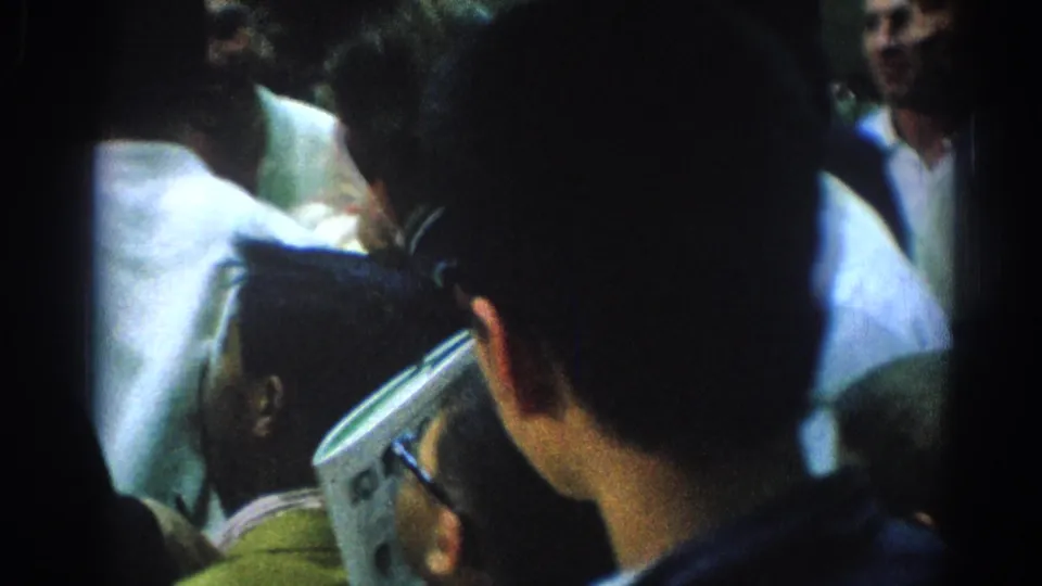 A low-light 8mm frame from San Gabriel showing attendees packed together, with one person holding a folded paper near the camera.