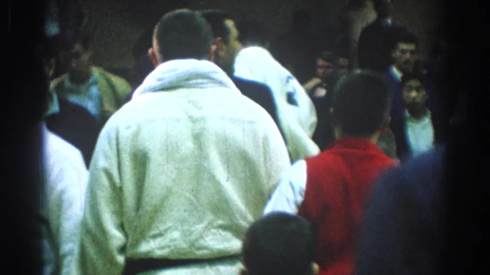 1966 San Gabriel crowd frame with a man in a light garment from behind, surrounded by attendees in a dim indoor space.