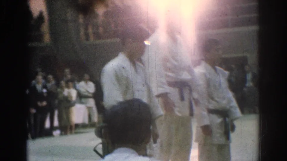 1966 San Gabriel competition ceremony view with a martial artist in a white gi, an official nearby, spectators behind them, and strong film flare.