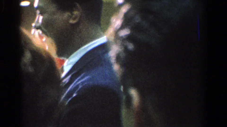 1966 San Gabriel home-movie frame showing spectators close to the camera and a suited man near an indoor martial arts event.
